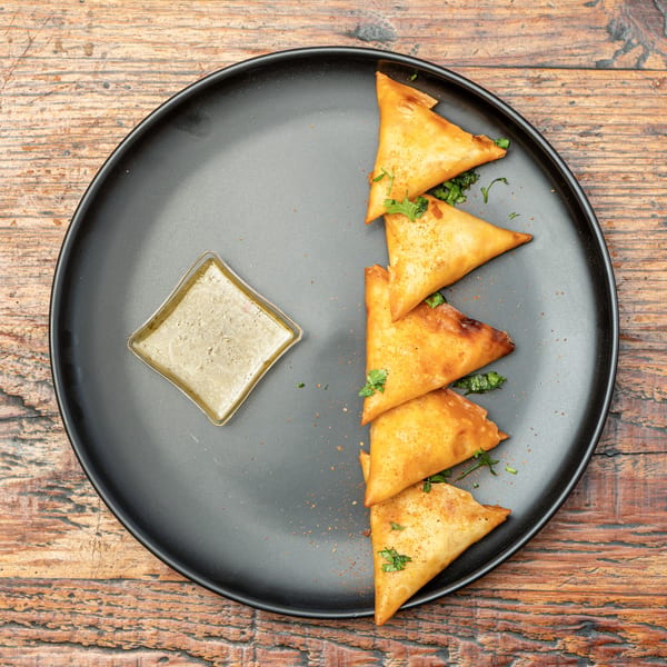 Crispy golden samosas with fresh herbs and a creamy dipping sauce on a black plate.