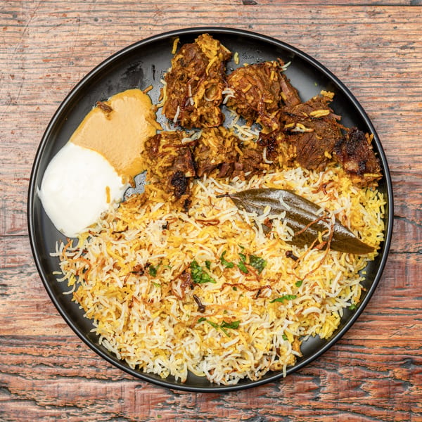 Biryani with spiced meat, fragrant rice, raita, and gravy on a black plate.