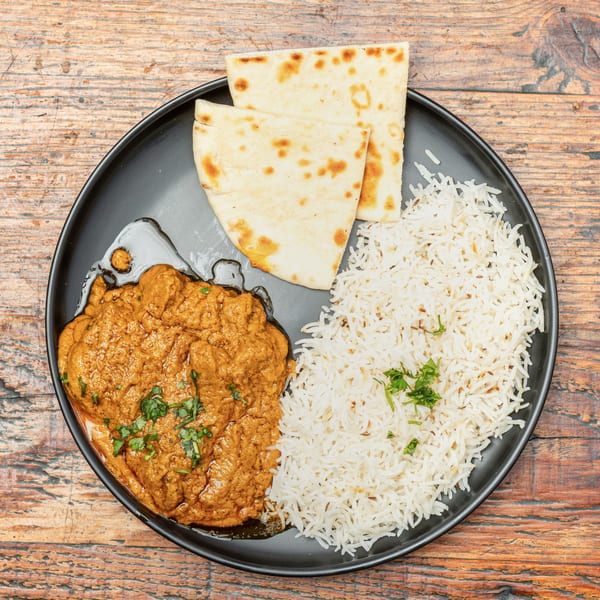 Indian curry with rice and naan bread on a black plate, set on a wooden table.