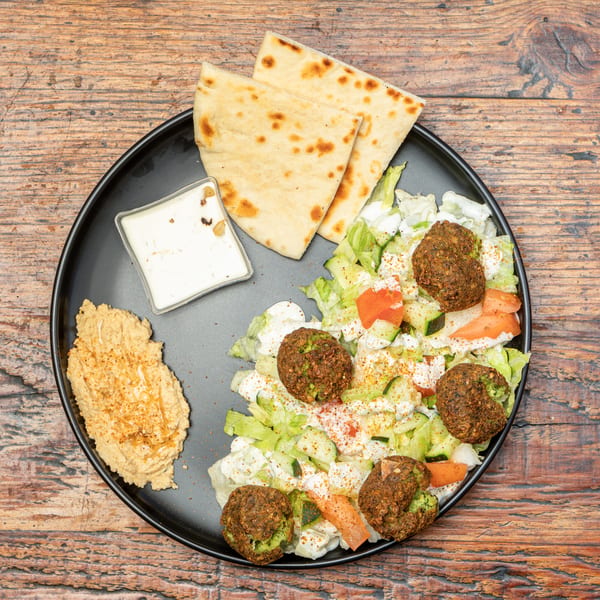 Falafel, fresh salad, hummus, pita bread, and white sauce on a black plate over a wooden table.