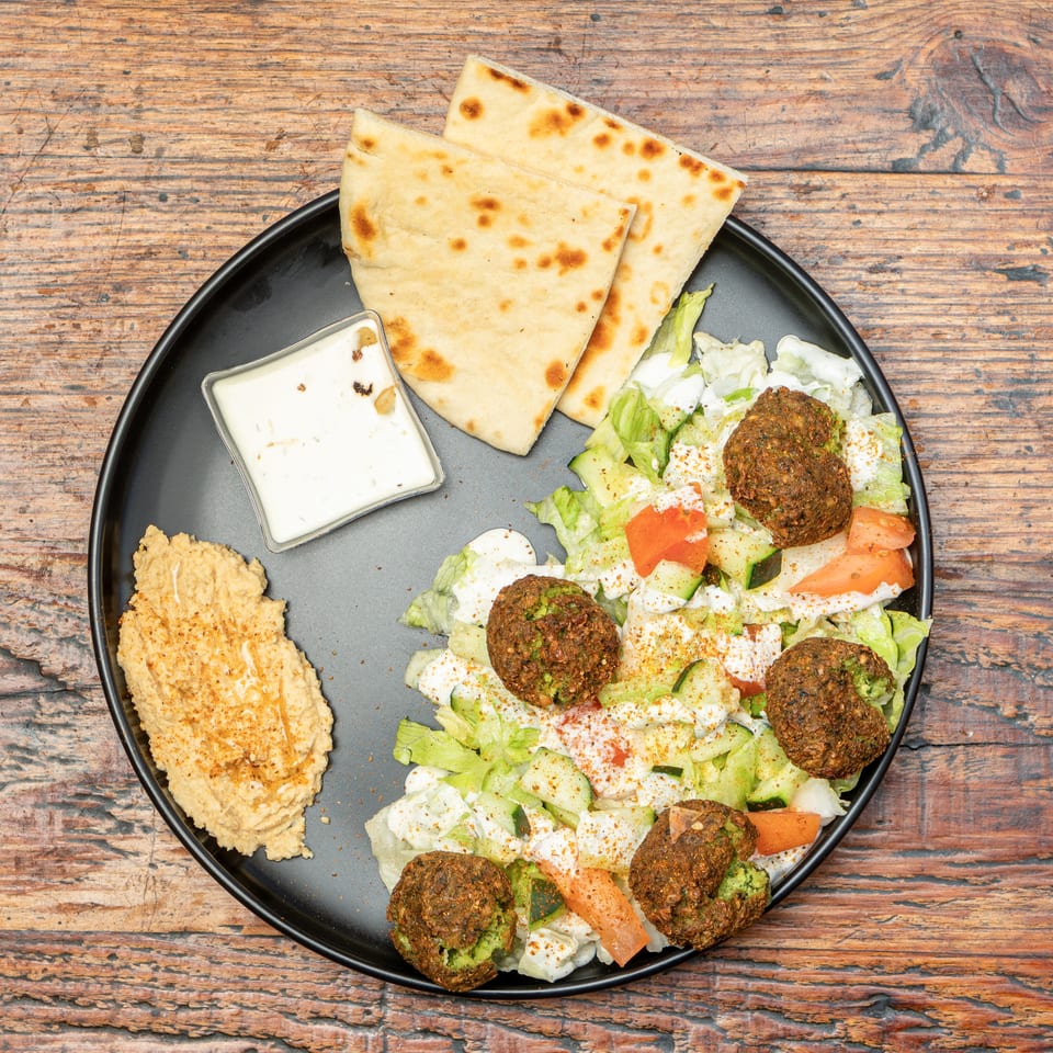 Falafel, fresh salad, hummus, pita bread, and white sauce on a black plate over a wooden table.