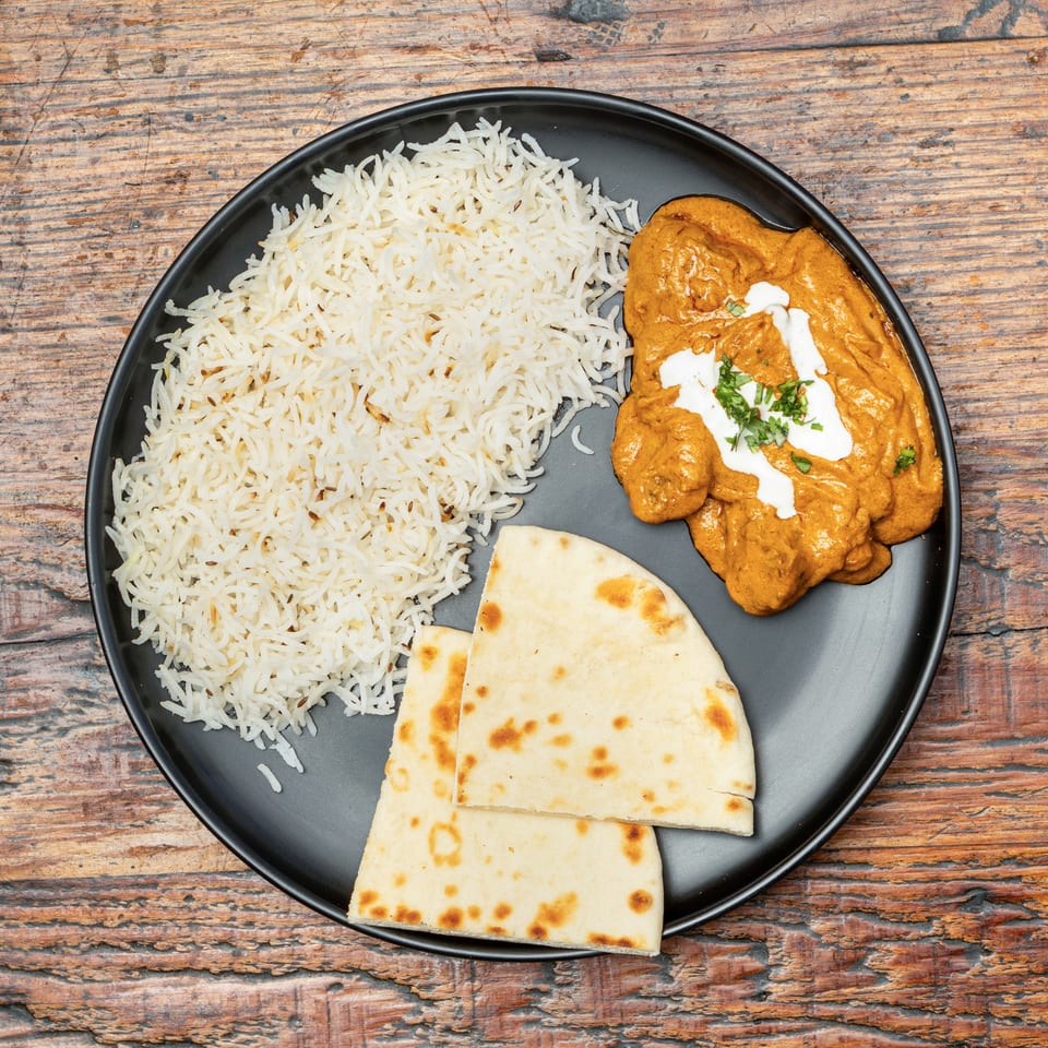A black plate holds basmati rice, creamy orange curry with herbs, and two triangular naan pieces.