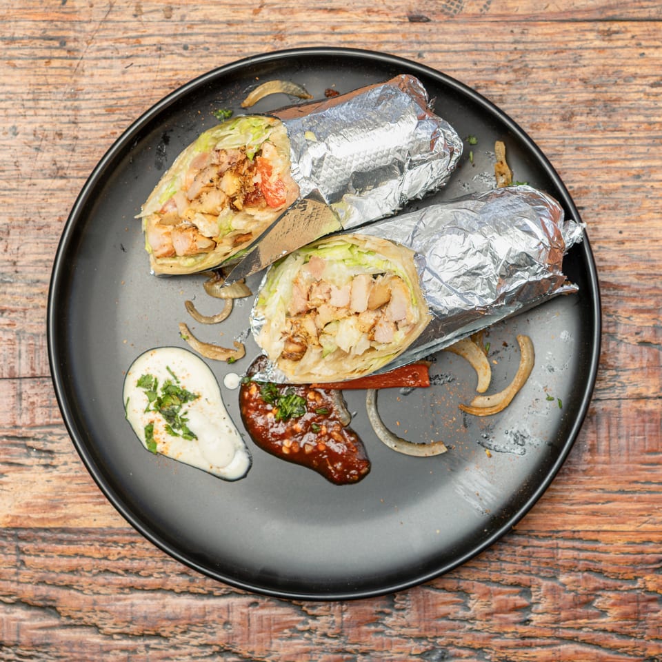 Two chicken wraps, partially unwrapped from foil, on a black plate with sauces and onions on a wooden table.